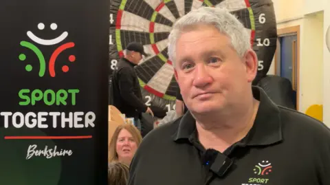 Craig Hunter is wearing a black polo shirt. He has short grey hair. He is standing in front of a black banner that says Sport Together Berkshire.