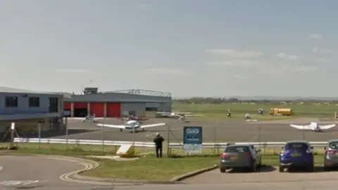 Google The run-way of Gloucestershire Airport with a car park in front and blue an black car parked inside.