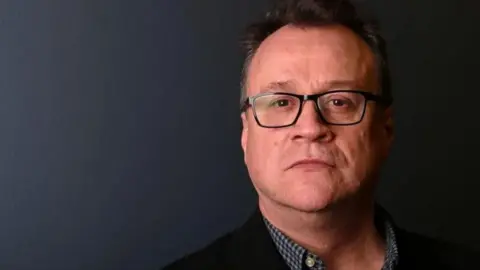 Getty Images Russell T Davies looking at the camera. It is a head and shoulder shot, he has a black jacket, under which he has a blue and white checked shirt with the collar undone. He is wearing black glasses. 