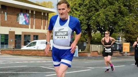 A man in blue running gear with a white bib, looks at the camera as he runs past on a road with other runners behind him.