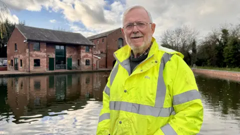 Bernie Jones stands near the canal basin with the restored brick warehouse building behind him. He has grey hair and is wearing a hi-vis jacket.