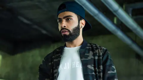 Raxstar The rapper Raxstar staring into the distance with a focused and stern expression. He is wearing a navy cap backwards on top of his head and has a dark beard. He has a cream jumper on with a camouflage jacket over the top of it.