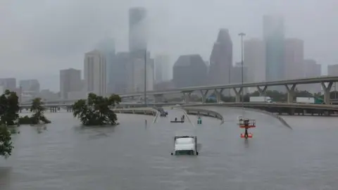 Reuters Houston skyline in the background, covered in cloud. In the foreground is a highway running into a large flooded area. A lorry, facing the camera, is partly submerged. There are a few trees on the left-hand side.