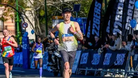 2026 Zurich Maraton de Sevilla Logan running the Seville Marathon in February 2026 as crowds line the streets with two other competitors behind. He's running in a yellow vest, grey shorts and black cap. Fans applaud the runners going by.