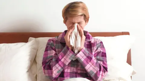A person sneezing into a tissue, she is wearing a pink and purple check pyjama top. She has light hair.