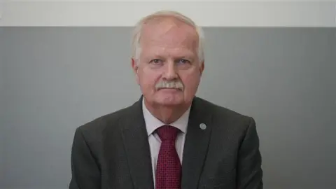 Jamie Niblock/BBC Robin Pivett wearing a suit, shirt and red tie. He has grey hair and a moustache and is looking directly at the camera with a serious expression.