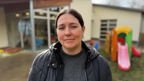 Steve Hubbard/BBC Karen smiling with lips closed into camera. She has dark hair worn in a ponytail with hooped earrings. She's wearing a grey sweater and a dark quilted coat with a hood and a zip front. She's outside in a playground setting with a multi-coloured children's slide over her left shoulder.   