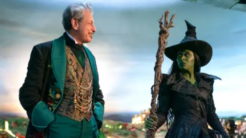 UNIVERSAL STUDIOS Jeff Goldblum as the Wizard in the Wicked movie wears an elaborate green suit and looking at Cynthia Erivo as Elphaba. She has green skin and wears a black witch's outfit.