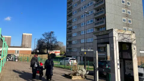 Two women in dark clothes walk with their backs to the camera holding the hands of a boy, who is wearing a red hat. The shell of a phone box is seen to the right and high-rise tower blocks to the right and in the distance.