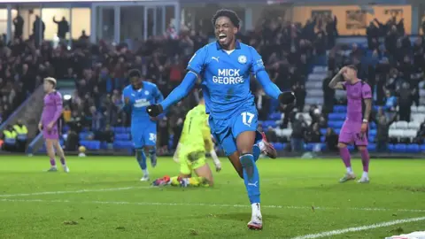 Getty Images Kyrell Lisbie cheering and running across the pitch at the Weston Homes Stadium in Peterborough. Behind him are disappointed Wigan players in a mauve away kit. 