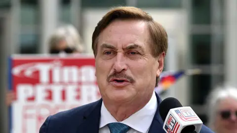 Mike Lindell has a mustache and speaks into a microphone while wearing a blue suit