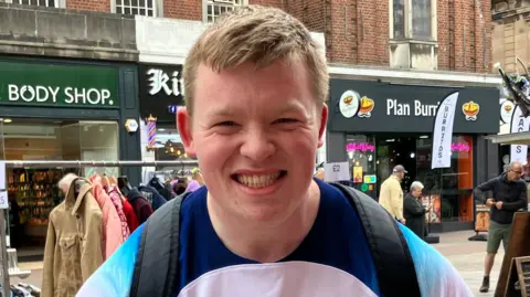 Aaron Smith is smiling at the camera wearing a white and blue England football jersey. He is standing in Southend high street with shops behind him.