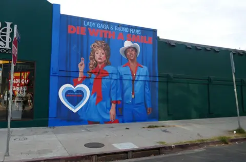 Alamy Um mural de arte em outdoor representando a capa do álbum do novo single de Lady Gaga e Bruno Mars, 'Die With A Smile'. Na arte, os dois usam ternos azuis de visual vintage com golas vermelhas sobre fundo azul. Também visíveis no quadro estão um sinal de pare, uma calçada, uma estrada e um céu.