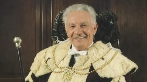 A smiling grey-haired man in robes with a fur-lined collar and a ceremonial chain on top. He is wearing a white bow tie and carrying a mace. There is a panelled wood background behind him.