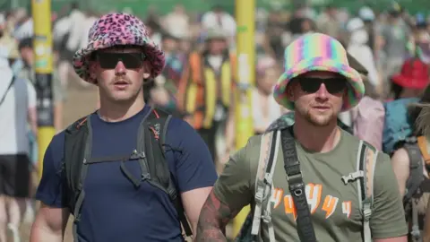 Two men wearing colourful bucket hats and carrying backpacks are walking towards through a crowd.