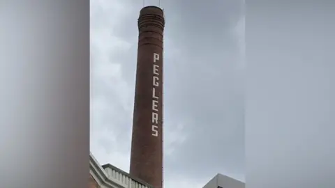 South Yorkshire Heritage List A thin red brick factory chimney stands against the skyline. Written horizontally down in white text is the word Peglers. 