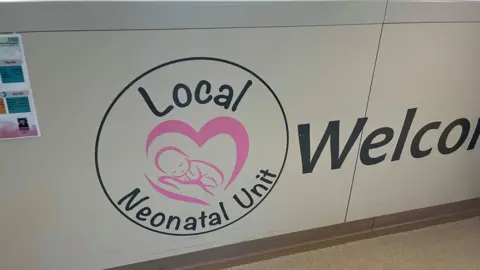 Ollie Conopo/BBC A white sign with a black circle which says Local Neonatal Unit, with a baby in a heart in the centre. The word "Welcome" appears in black lettering to the right. There is a notice attached to the sign to the left.