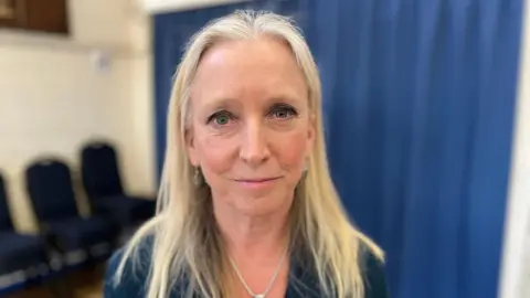 MP Roz Savage smiling, wearing a necklace and blue top. Blurred behind her is a room with a blue curtain and stacked chairs