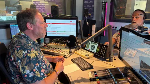 Amy Holmes/BBC Two people, radio presenter Olly Mann on the left, and the Mayor of Bedford on the right are pictured conducting an interview in a professional radio studio surrounded by microphones, computer screens, and audio mixing equipment.