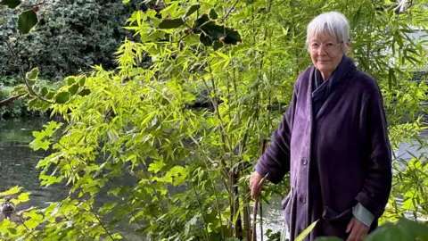 Eric Johnson/BBC A grey-haired woman in a purple coat stands on the edge of the canal from her garden.