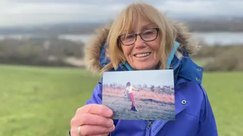 A woman in a thick blue winter jacket and wind-swept blonde hair holds up a picture of a younger woman skiing on grass.