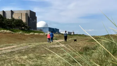 Three people walking on beach path in front of Sizewell A and B power stations on a sunny day with some clouds in the sky. There is a small black dog walking alongside them.