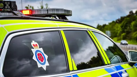 A police car is parked facing a road. It has a sticker on the back window which reads Lancashire Constabulary. A blue sky and some trees can be seen in the distance over a road. 