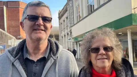 Andrew Turner/BBC Nigel Peart has grey hair and has tinted spectacles. He is wearing a grey hoodie and blue polo shirt beneath. His wife, to the right of the image, also has tinted spectacles. She has greying long hair, and is wearing a black faux-fur lined jacket and a red polo neck jersey.