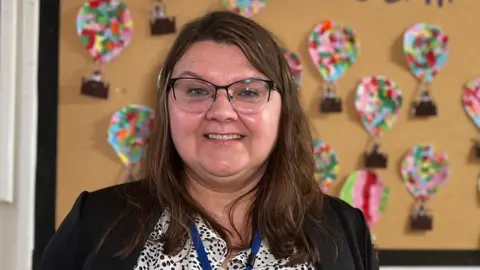 Monika is standing in a classroom in front of a bulletin board decorated with colorful hot air balloon cutouts. The board has the text “Flying High in Year 1” written at the top. She is wearing a patterned blouse, a black cardigan, and a visible name badge that reads “Monika.”