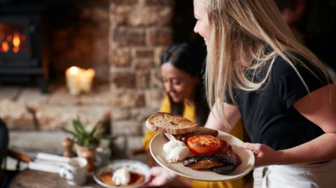 Generic image of hospitality worker bringing a full English breakfast to customers at a table in a traditional pub