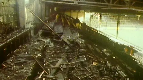 A burnt out football stand, showing fallen wood and charred remains.