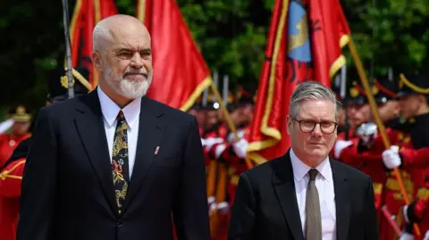 PA Sir Keir Starmer arrives for bilateral meeting with Prime Minister Edi Ramat at the Kryeministria, Tirana, Albania, a bearded man who is much taller than him. Both are wearing dark suits and ties and there are red ceremonial flags in the background