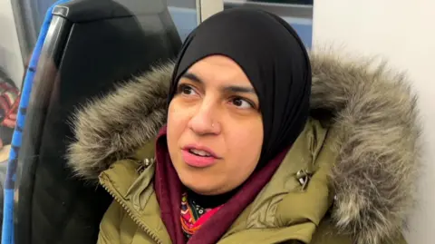 Sarah wearing a black headscarf and a khaki green coat with a fur lined hood, wearing a maroon scarf while sitting on a train seat.