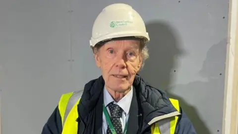 Lawrence is wearing a white hard hat. He has a blue shirt and tie with a padded blue jacket over the top. He also has a high vis yellow jacket on and a green lanyard.