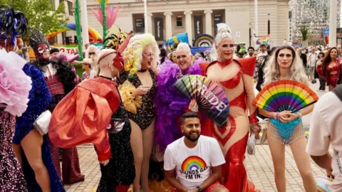 A group of drag queens who are taking part in a city's Pride parade. They are all smiling and standing together in a semi-circle. A man in crouched down in front of them. He is wearing a white top with a rainbow on it.