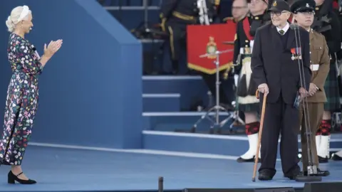 PA Media/ Andrew Matthews Stage with band members in background - man with walking stick to right wearing navy suit adorned with medals - Dame Helen Mirren in floral dress is clapping towards the man.