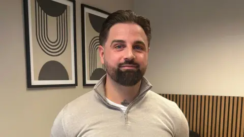 A man with a beard is wearing a beige jumper with a zip and has a tattoo visible at the base of his neck