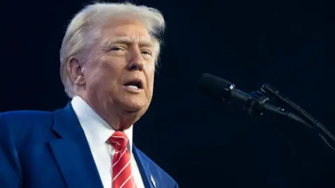 Donald Trump talking into a microphone. He has white hair swept back over his head and is wearing a blue suit with a white shirt and red and white striped tie.