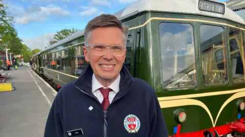 Phil Harrison/BBC A man in glasses and a blue fleece stands besides a green train with cream and red markings. 