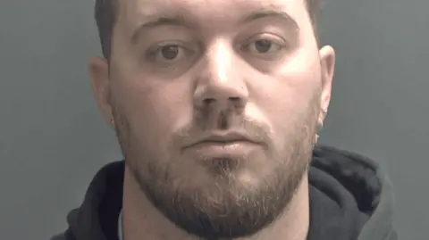 Christopher Rudd is staring at the camera and has a light brown and beard and earrings. He is wearing a dark hoodie and is standing against a plain grey background.