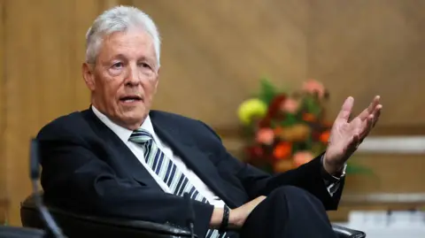 Press Eye Peter Robinson has short grey hair and is wearing a dark suit, with a blue stripped tie and a white shirt. He is gesturing with his left hand. He is sitting in a chair in a room with wooden panelling and vase of flowers in the background.