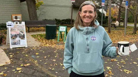 Woman in light blue sweatshirt holding mug stands in front of building, signs and benches 