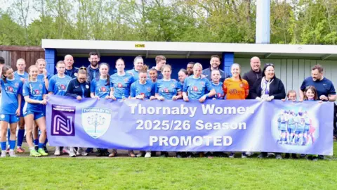 Sam Kirkup Thornaby Women are lined up in blue football kits behind a banner reading "Thornaby Women 2025/26 Season PROMOTED".