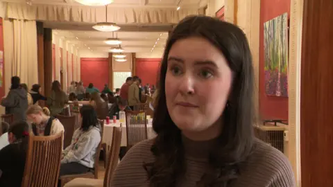 BBC A girl with long thick brown hair and a brown top. She has a neutral expression and stands in front of the camera. Behind her tables with groups of people sitting at them can be seen