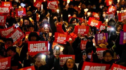 Reuters A crowd of protesters fills the frame, holding electric candles and red placards. One man in focus in the centre holds a gloved hand to his mouth as he yells