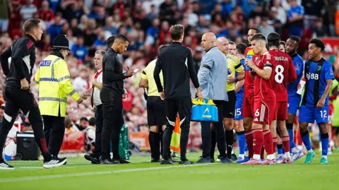 PA Media A crowd of Liverpool and Bournemouth players, including Semenyo, stand at the touchline alongside the refereeing team and Liverpool boss Arne Slot during the match pause. A policeman in a helmet and yellow jacket is speaking to a club offical while another match official writes notes on a pad and another man carries a bag of drink bottles.