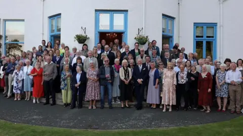 GOVERNMENT HOUSE A group of smartly dressed people standing in a bunch outside Government House for a photograph. The walls of the building are white and the door and window frames are painted light blue.