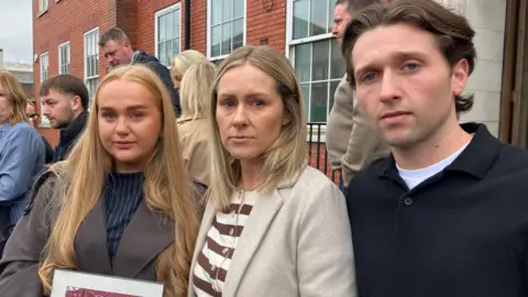 A woman with long dark blonde hair and a brown jacket holds back tears as she holds a photo of a woman with blonde hair cheering in a stadium. She is standing next to a woman in a cream and brown striped jumper and cream jacket with medium length light blonde hair. She is standing next to a man in a black jumper and brown hair. 