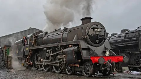 John Titlow Driver Mick Matthews whistles up, sitting inside the carriage, with fireman Alex Robinson standing beside him, as the black and red 75069 train is prepared for events in December.