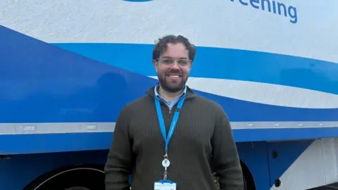Casper Lisle-Pourzyaie, Operations Manager for the lung cancer screening team in Coventry and Warwickshire stands in front of the mobile unit with white and blue markings on it. he has short dark hair and a short dark beard.